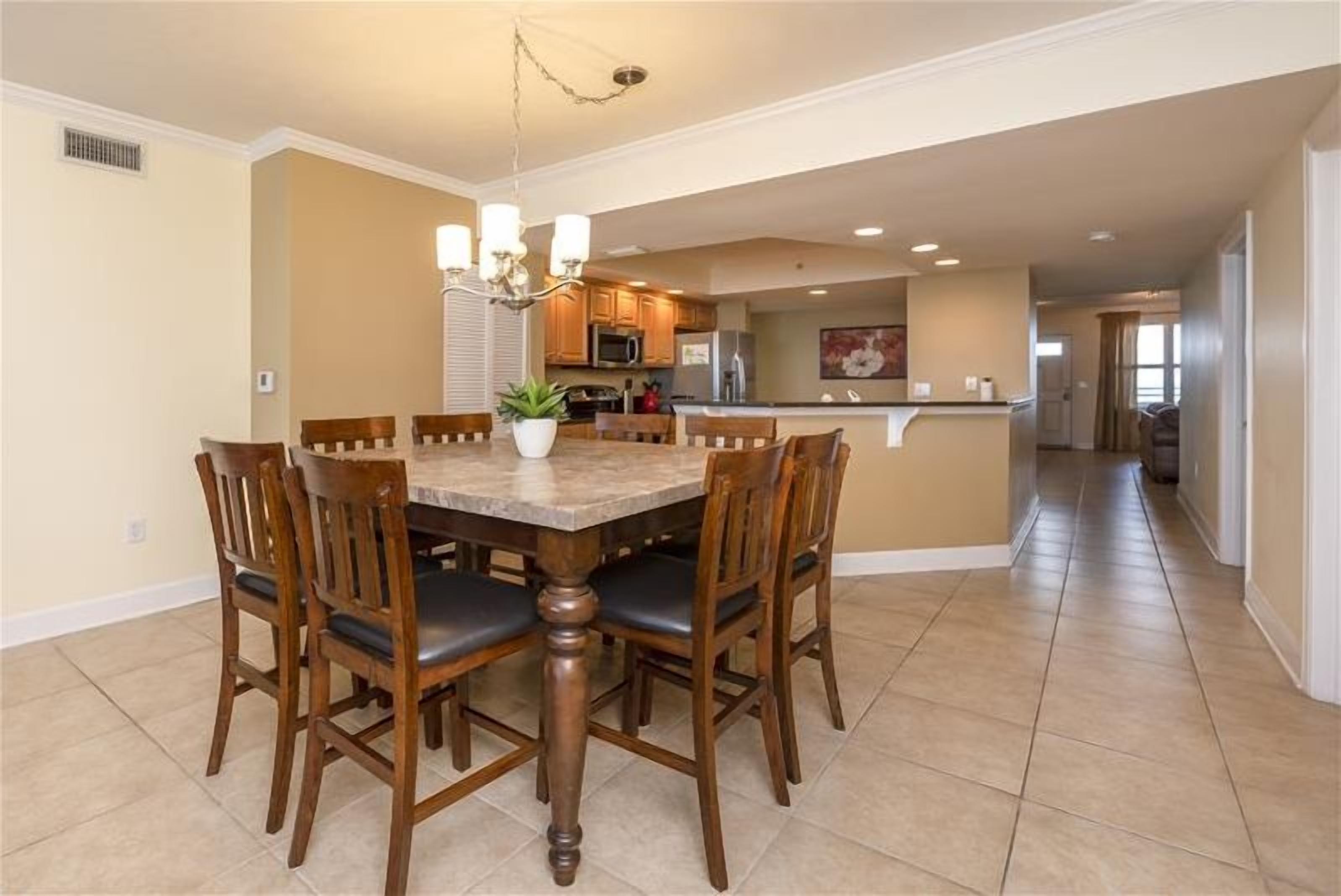 A dining area with a wooden table and chairs, a chandelier, and a kitchen in the background.