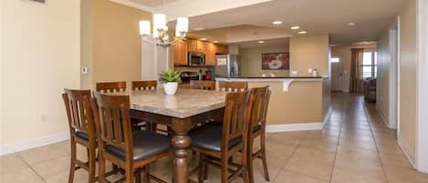 A dining area with a wooden table and chairs, a chandelier, and a kitchen in the background.