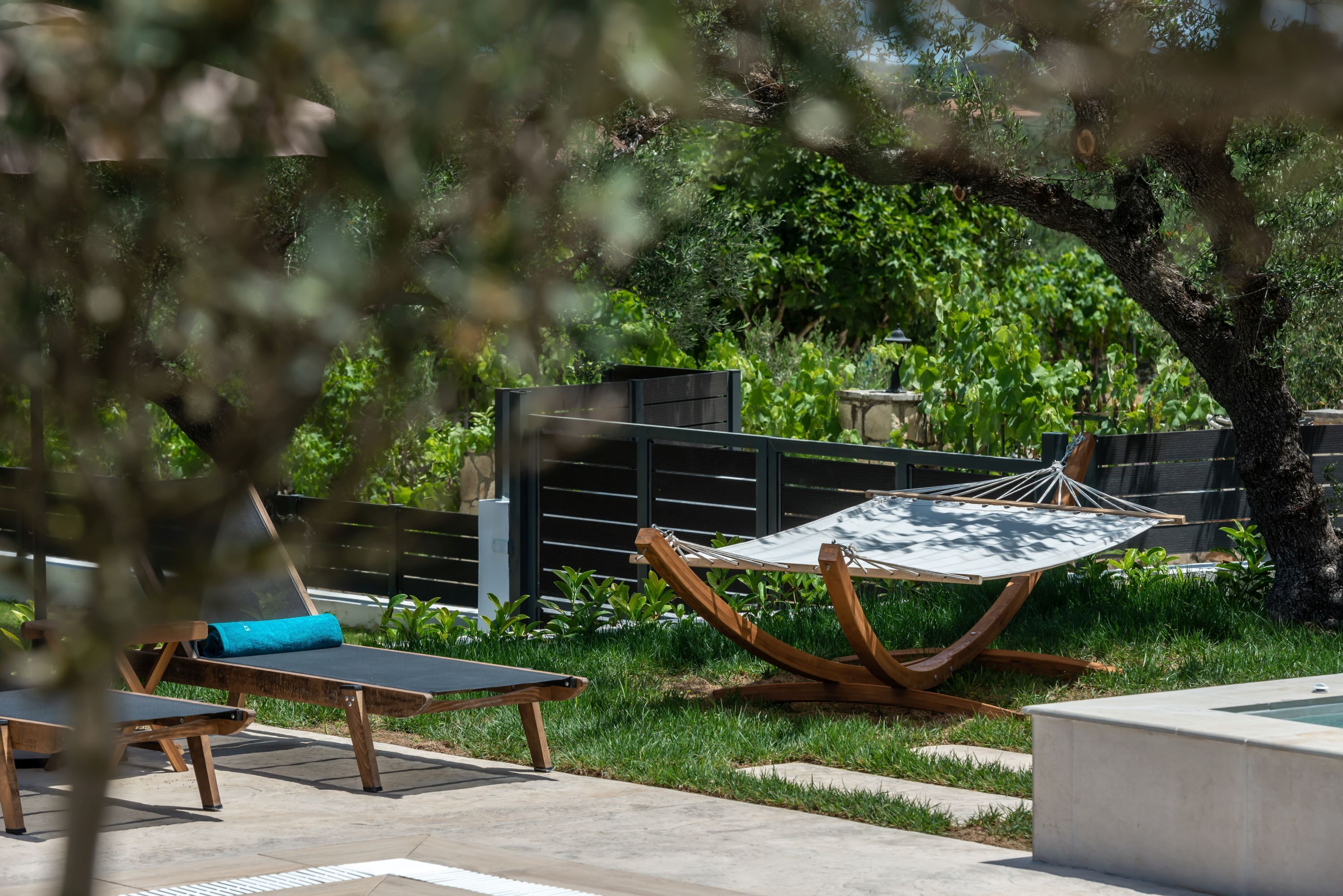 A hammock chair, a wooden lounge chair, and a small table in a garden area with a fence and greenery in the background.