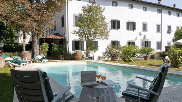 A poolside area with lounge chairs, a table with a book and glasses, and a building with shutters in the background. - 1800 Tuscan villa - walking distance from a pretty village (Fornoli)
