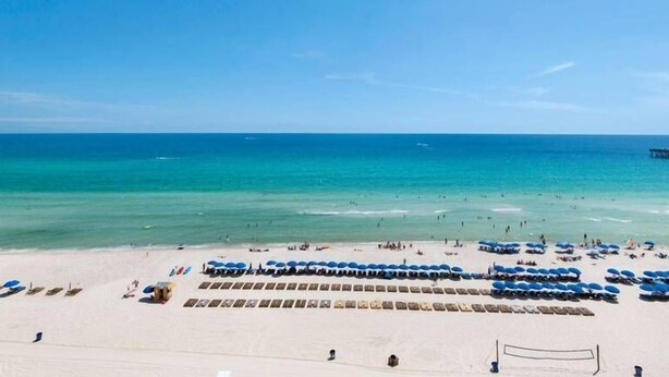Une plage bordée de rangées de parasols bleus et de chaises longues, un ciel bleu dégagé et une mer calme.