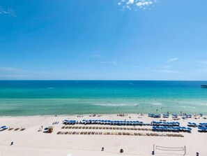 Una playa con hileras de sombrillas azules y tumbonas, cielo azul despejado y mar tranquilo.