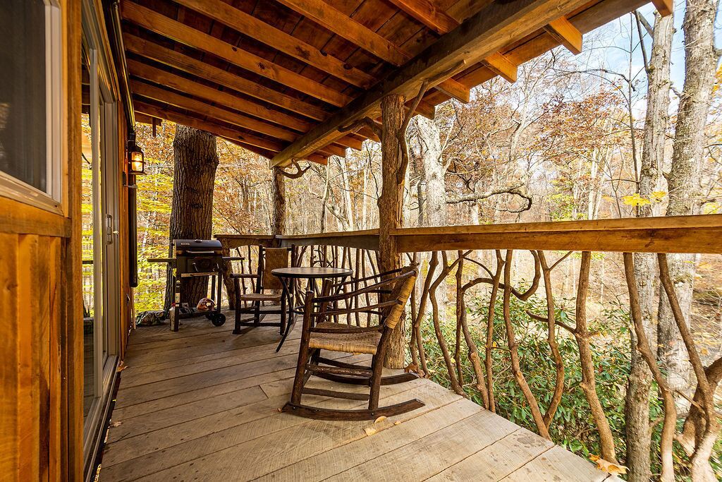 A wooden deck with a rustic wooden chair and a table, surrounded by trees.