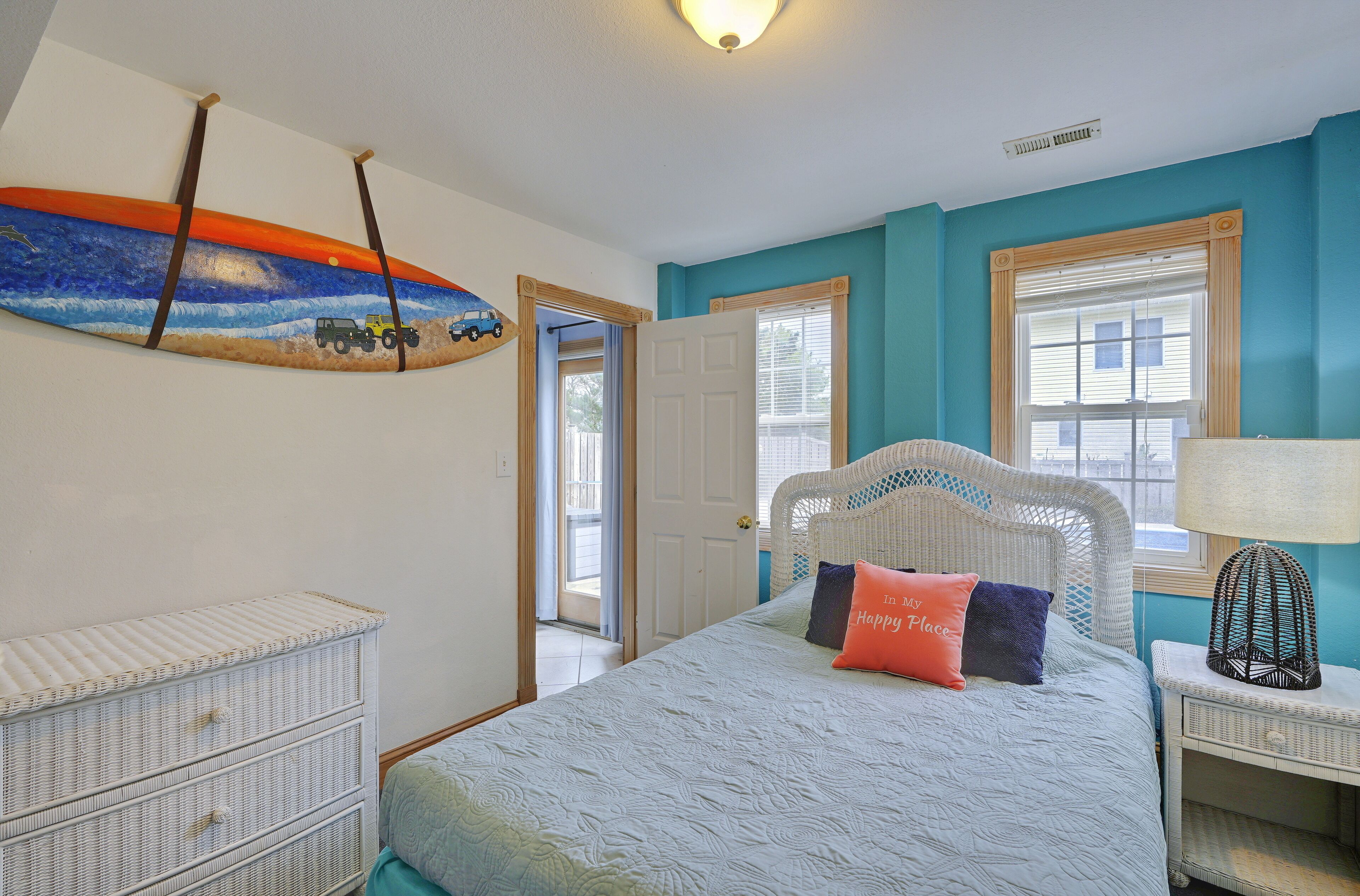 A bedroom with a bed, a dresser, a surfboard painting, and a window with blinds.