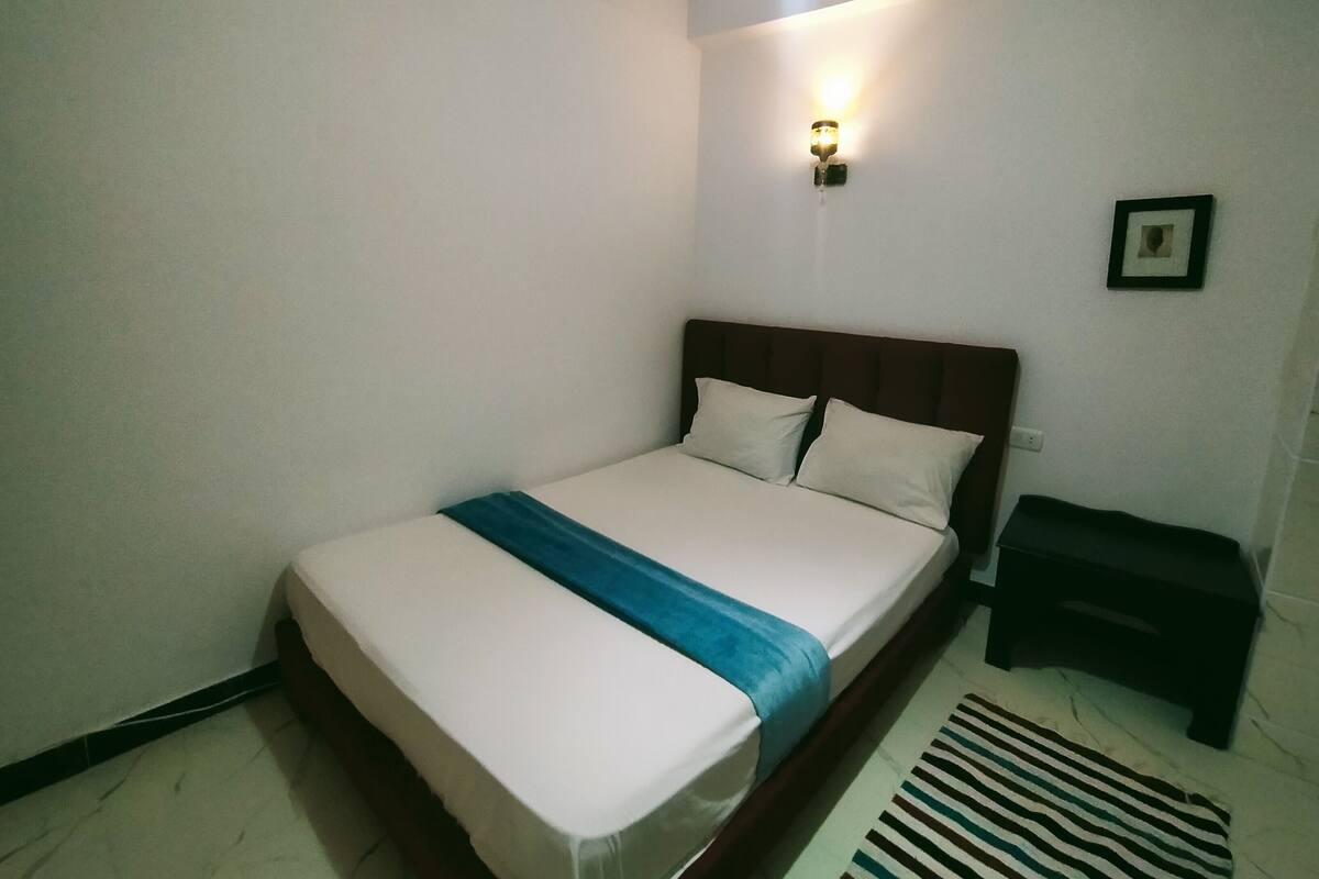 A single bed with a wooden headboard, white bedding, and a blue accent. A small bedside table and a framed picture are visible.