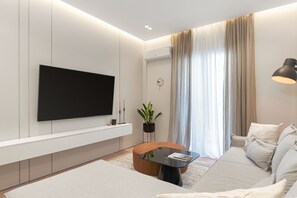A modern living room with a flat-screen TV mounted on the wall, a round coffee table, a sofa, and a large window with curtains. - Acropolis White Key Residence (Athens)