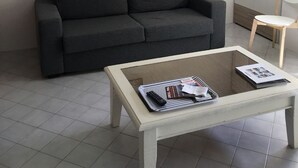 Uma sala de estar com um sofá cinza escuro, uma mesa de centro branca com revistas, e uma placa na parede com a palavra 'WELCOME'.