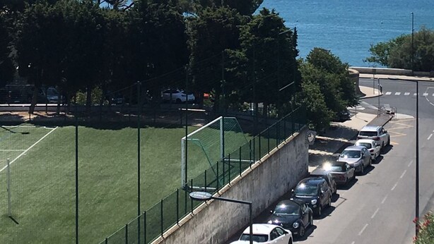 Um parque de estacionamento ao lado de um campo desportivo, com vista para o mar.