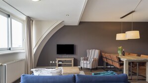 Een moderne woonkamer met een blauwe bank, een gestreepte fauteuil, een eettafel met stoelen, een flatscreen televisie en een groot raam.