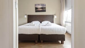 Een tweepersoonsbed met witte lakens en twee kussens, een donker hoofdbord, bedlampjes en een schilderij van een zonsondergang boven een meer aan de muur.