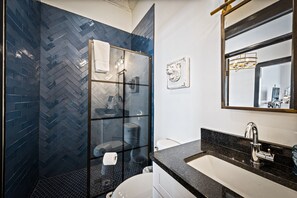 Bathroom features a herringbone tile pattern, glass-enclosed shower, white toilet, and large mirror with a gold frame.