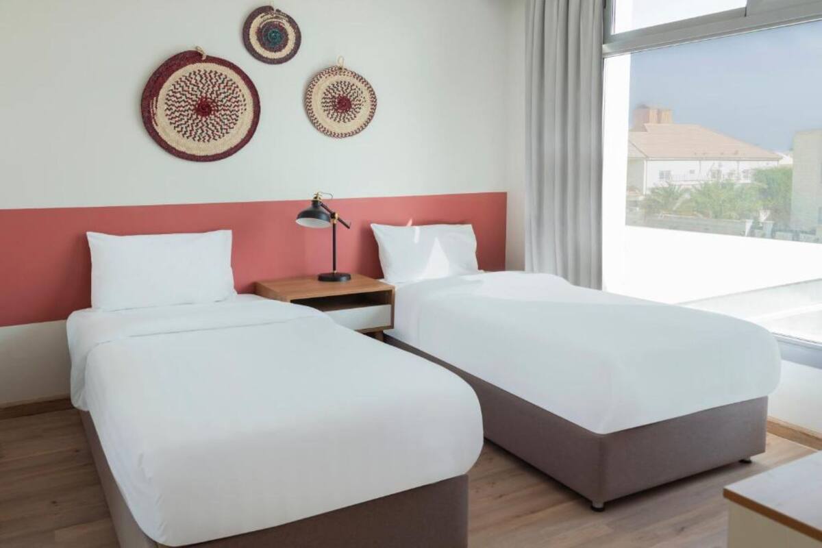 A hotel room with two beds, a nightstand, a lamp, and a view of the outside.
