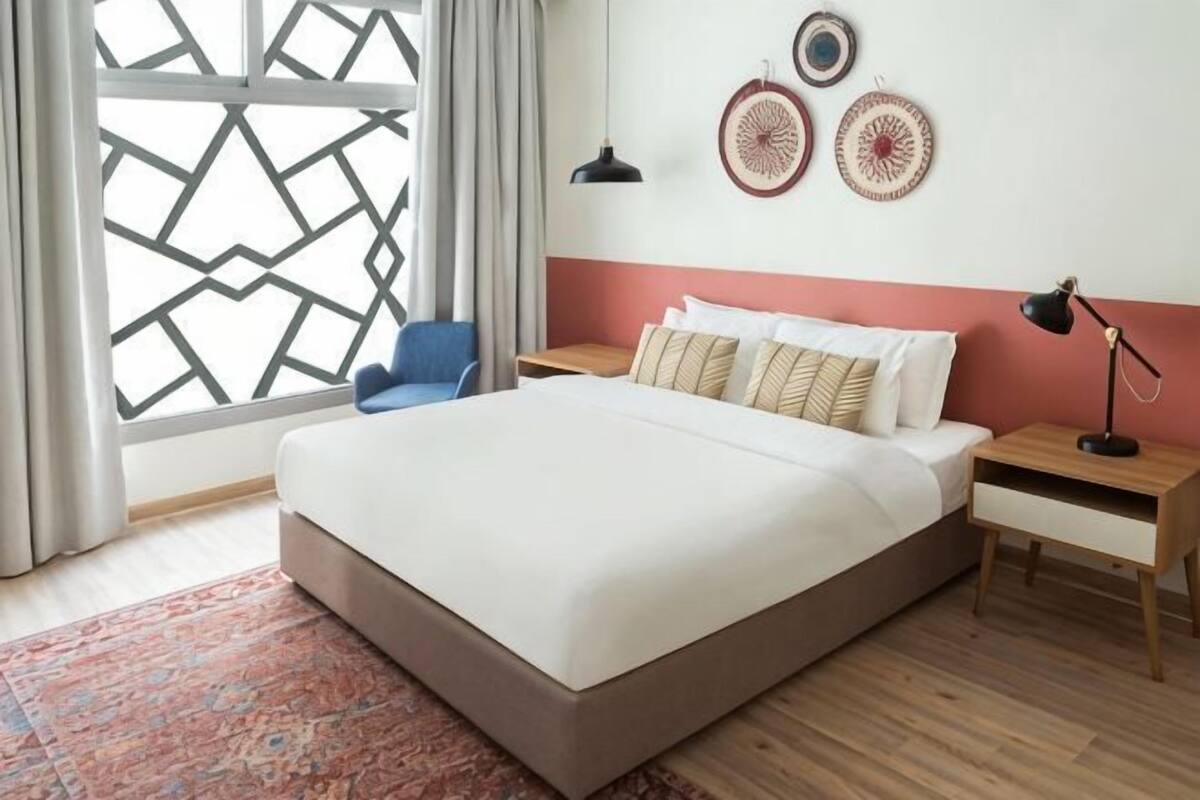 A bedroom with a large bed, a nightstand, a desk, a chair, and a window with a geometric pattern.