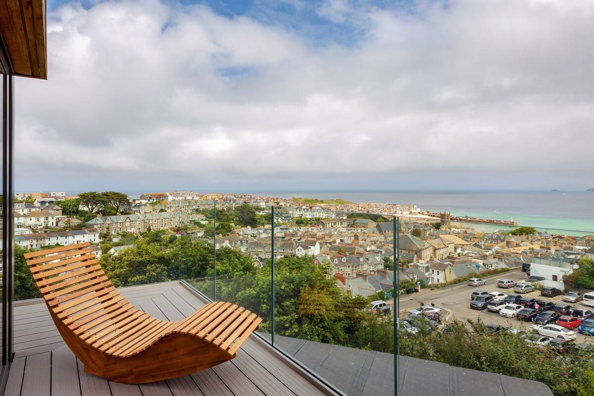 A wooden lounge chair on a balcony overlooking a coastal town with a view of the sea and buildings.