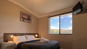 A bedroom with a bed, bedside lamps, a nightstand, a TV mounted on the wall, and a window with a view of the landscape.