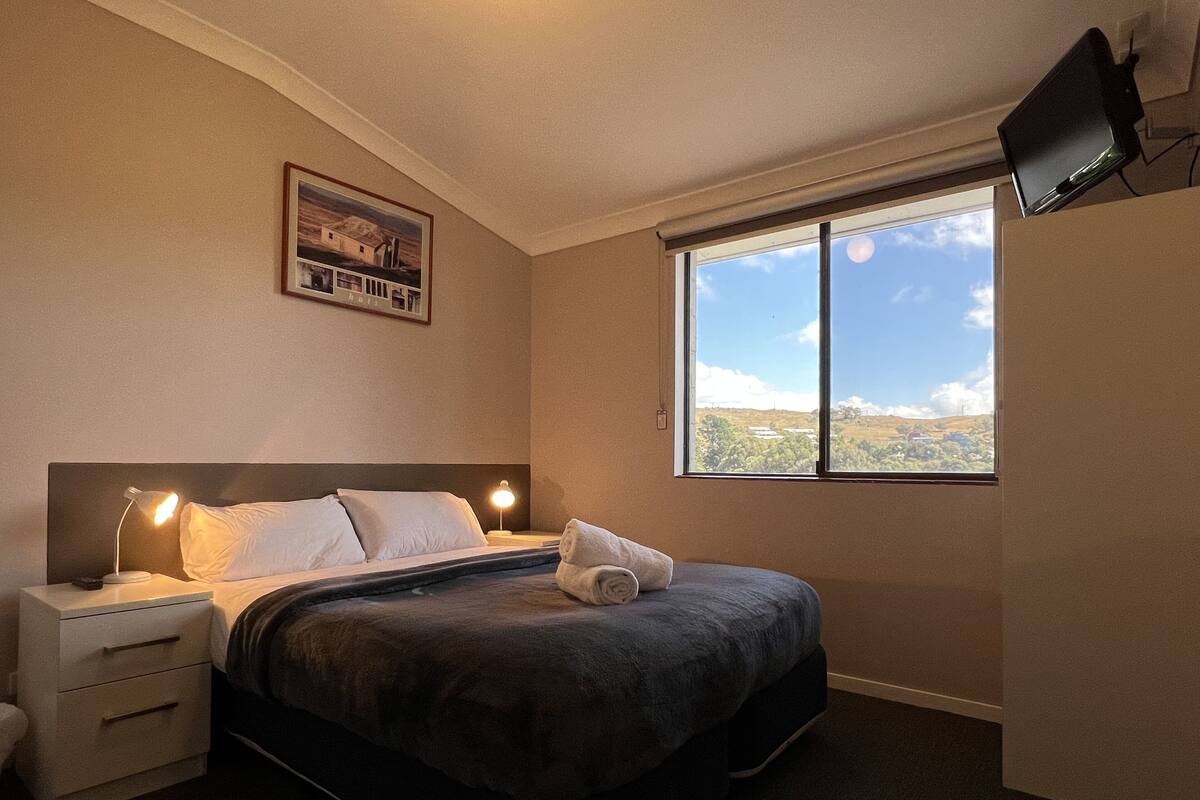 A bedroom with a bed, bedside lamps, a nightstand, a TV mounted on the wall, and a window with a view of the landscape.