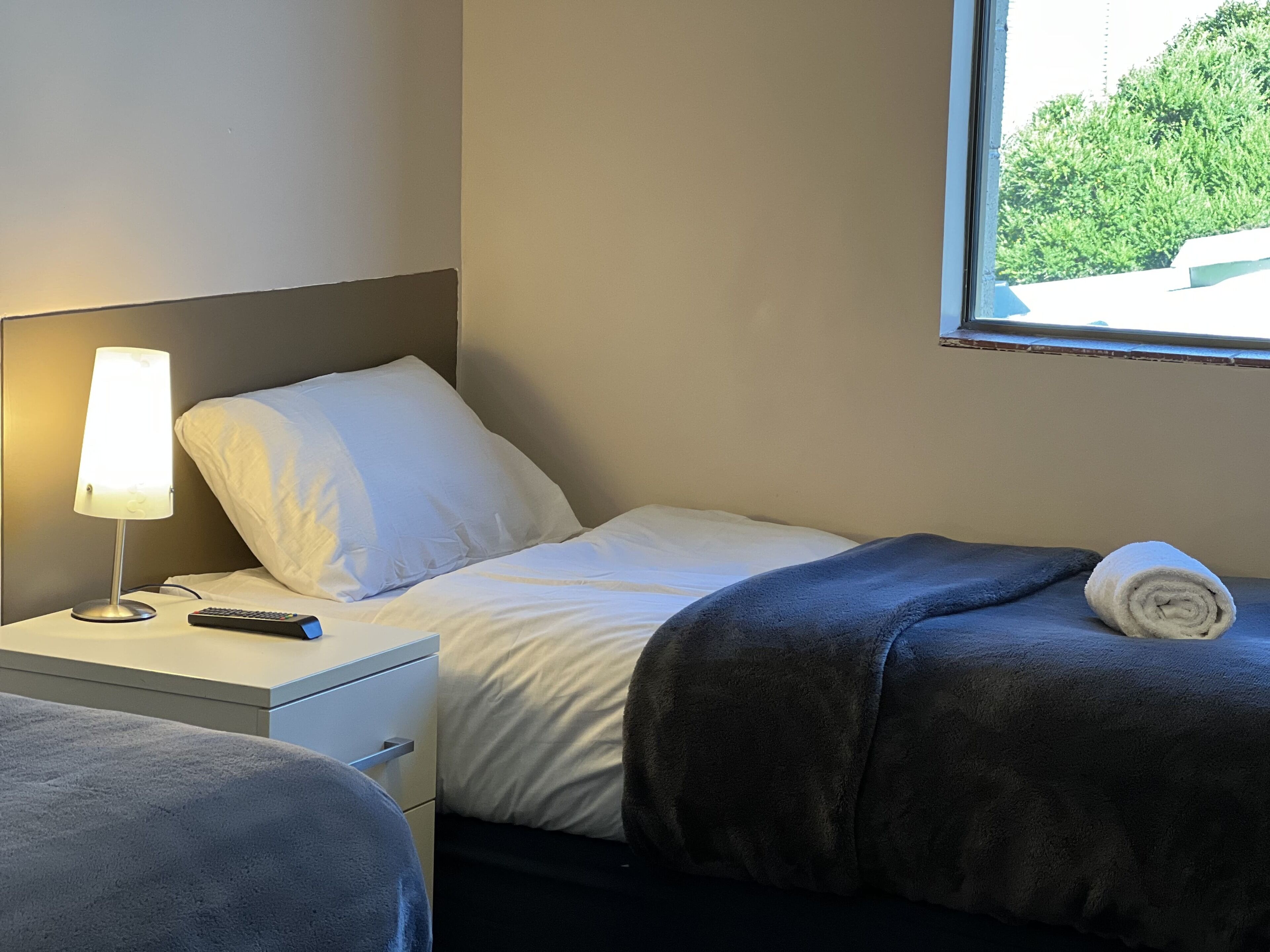 Una camera d'albergo con un letto, un comodino con una lampada, un telecomando e una finestra vista verde.