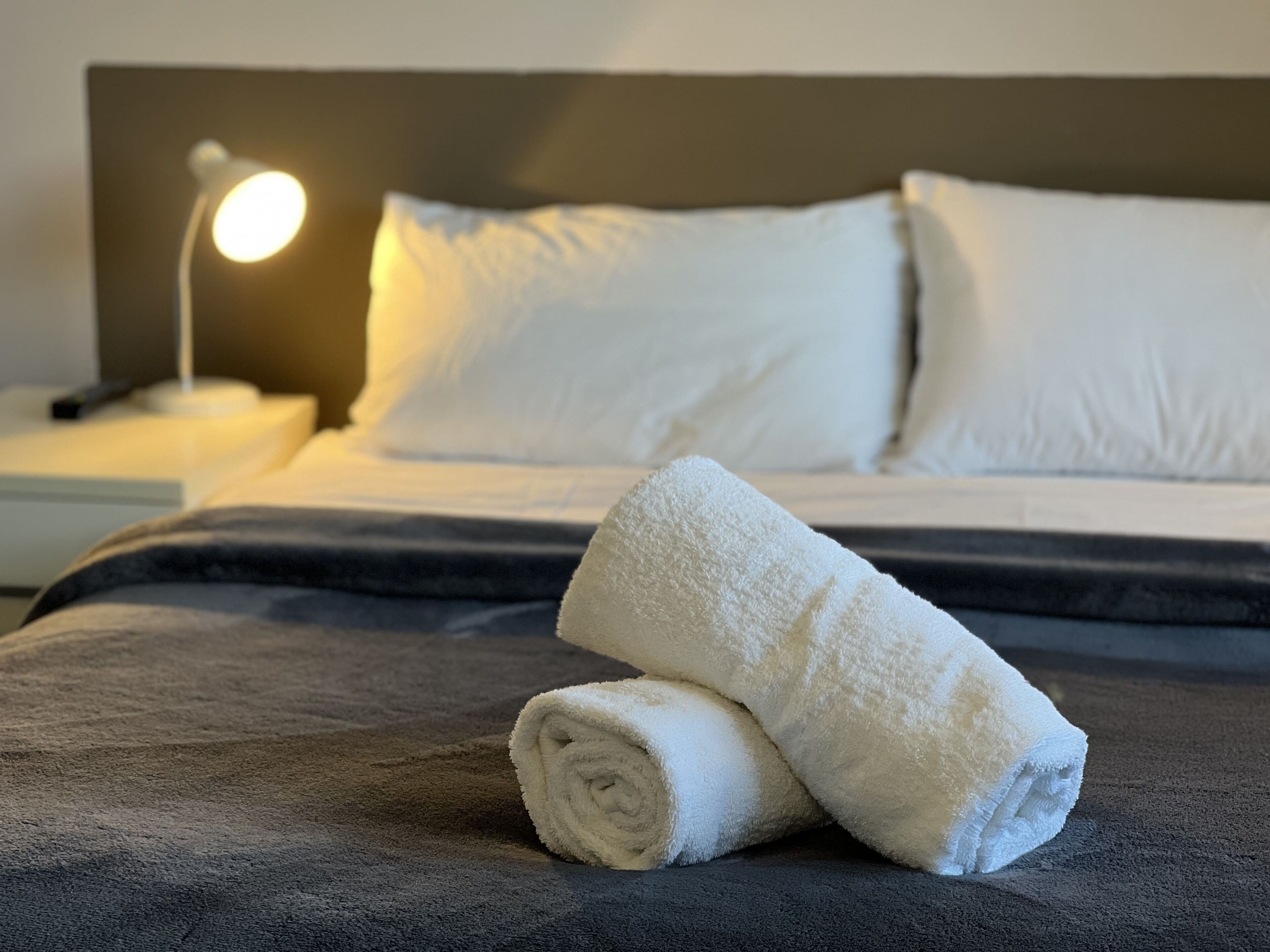 A neatly made bed with white linens and a rolled towel on a dark gray bedspread.