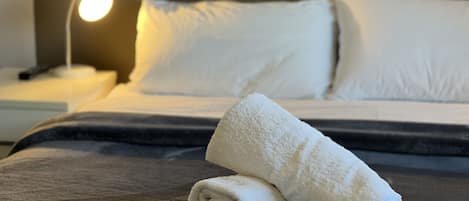 A neatly made bed with white linens and a rolled towel on a dark gray bedspread.