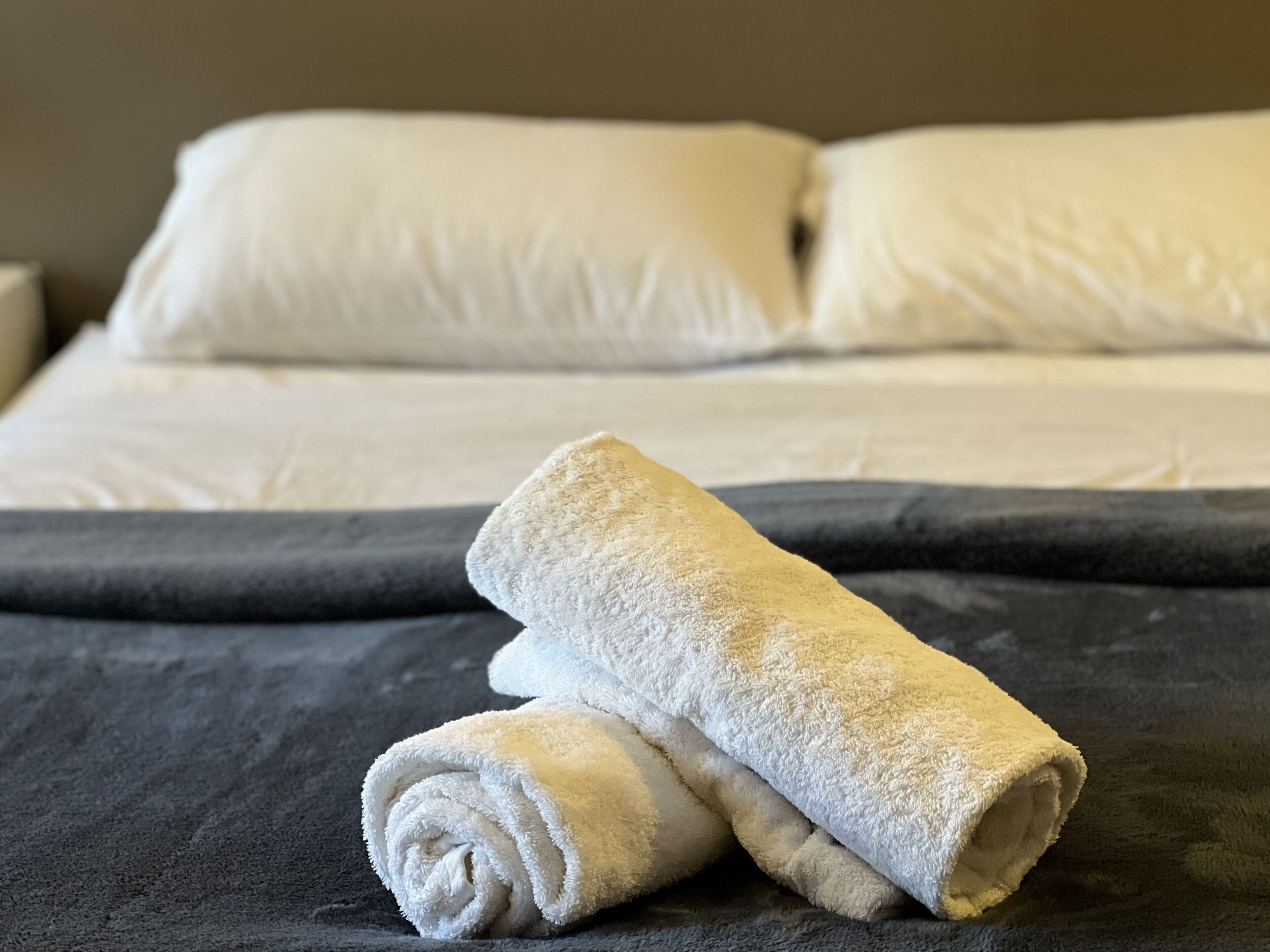 Una cama bien tendida con sábanas blancas y una toalla enrollada sobre un edredón oscuro.