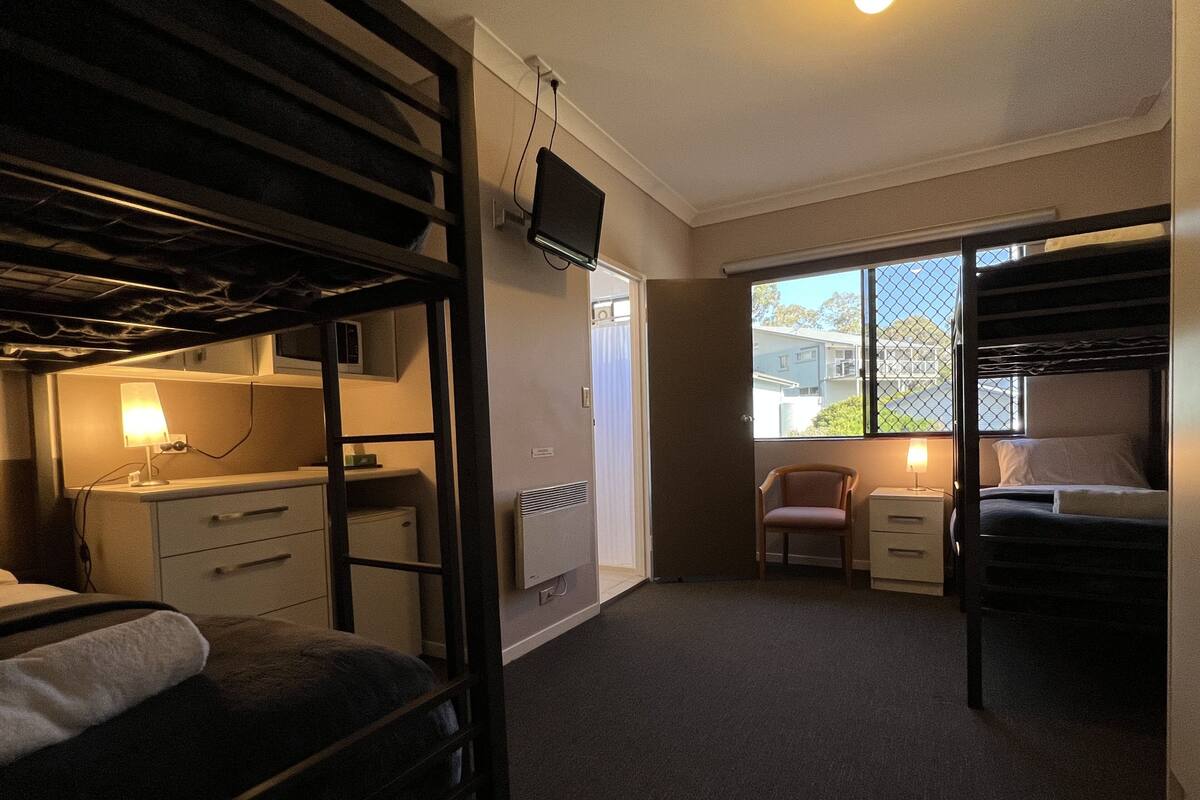 A compact hotel room with bunk beds, a desk with a lamp, a chair, and a window with a view of buildings.