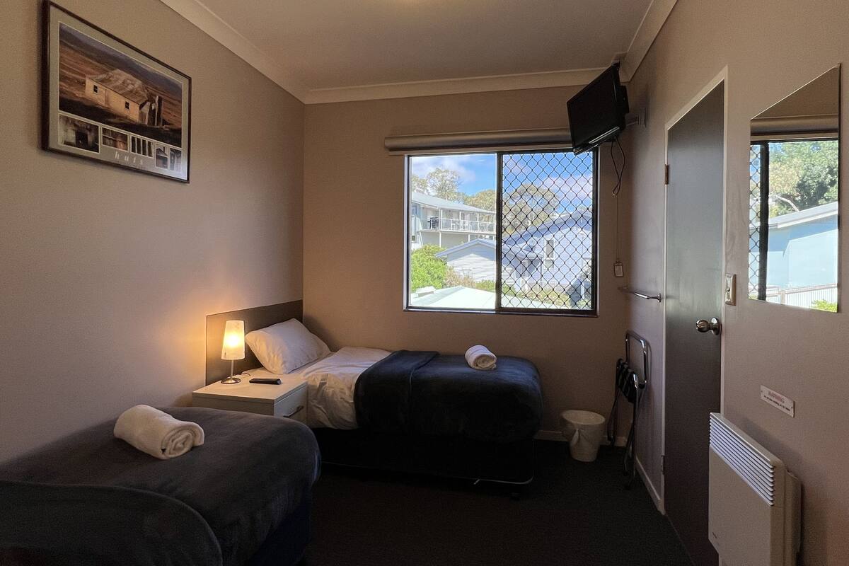A hotel room with two beds, a window with a view of buildings, a TV on the wall, and a door with a warning sign.