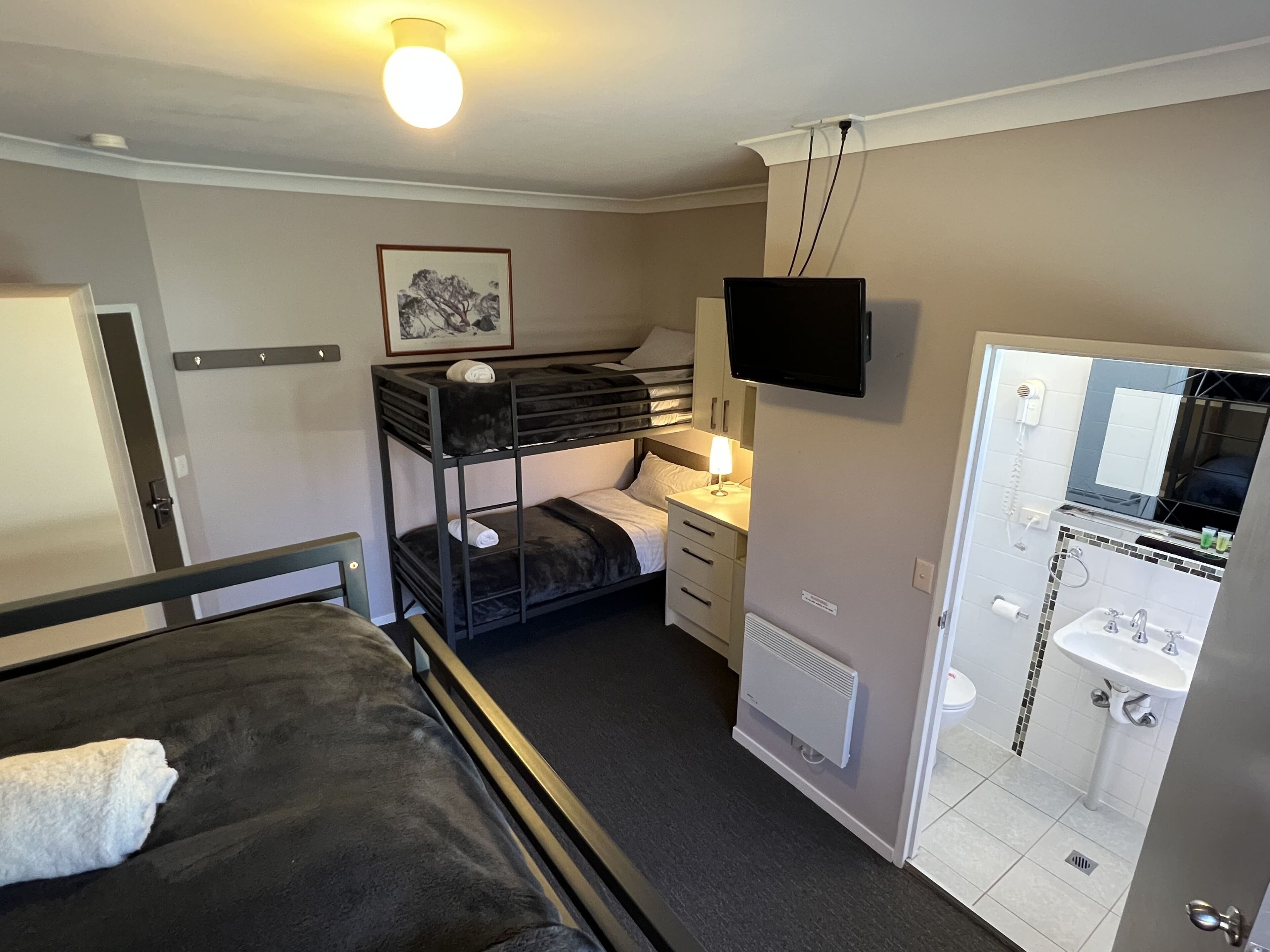 A compact hotel room with bunk beds, a desk, a TV, a bathroom with a sink and mirror, and a framed picture on the wall.