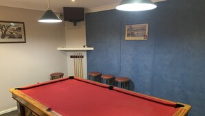 A pool table with a red felt surface, a wooden side, and a white cabinet with a speaker above it. There are three stools and a framed picture on the wall.
