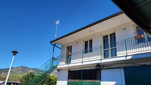 Um edifício de dois andares com varanda, escadaria e montanhas ao fundo.
