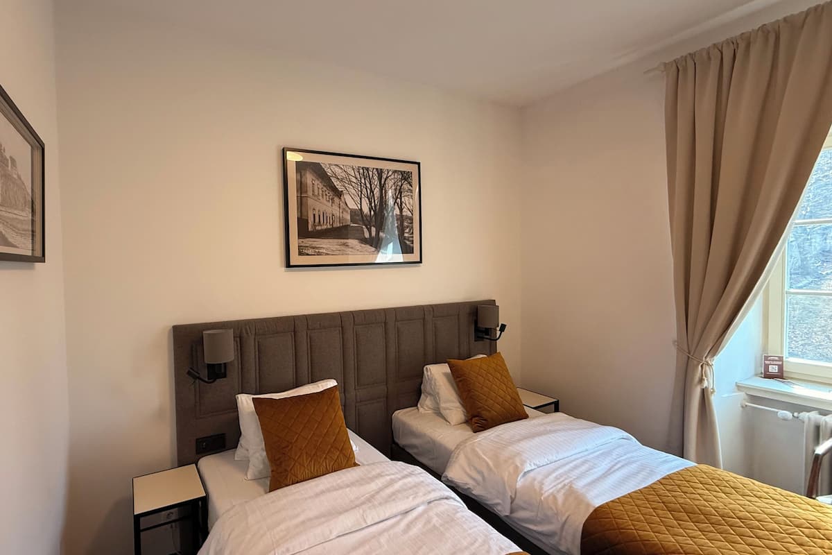 Habitación de hotel con dos camas, cabeceras, mesitas de noche, un cuadro enmarcado en la pared y una ventana con cortinas.