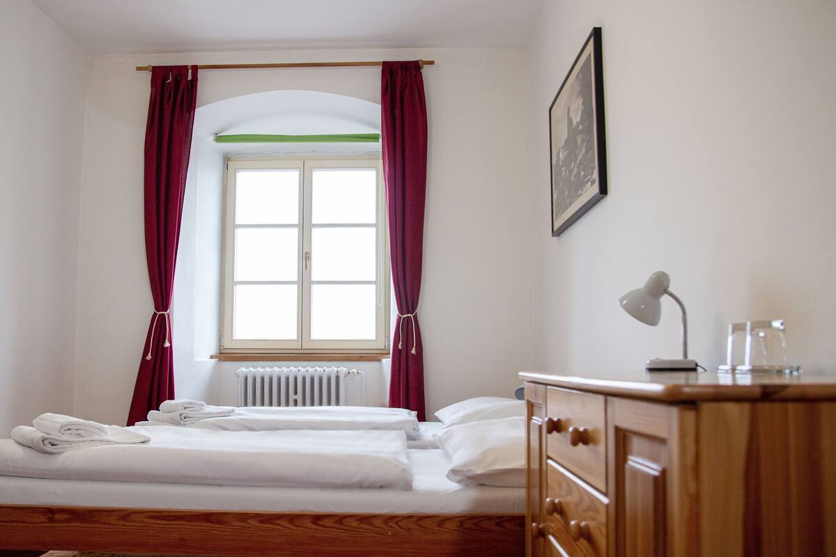 Una habitación con cama, cómoda de madera, escritorio con lámpara y un cuadro enmarcado en la pared.