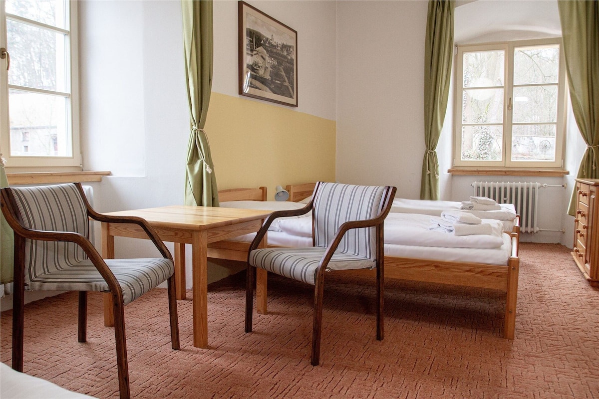 Una habitación con una cama de madera, dos sillas a rayas, una mesa de madera, un radiador y una ventana con cortinas.
