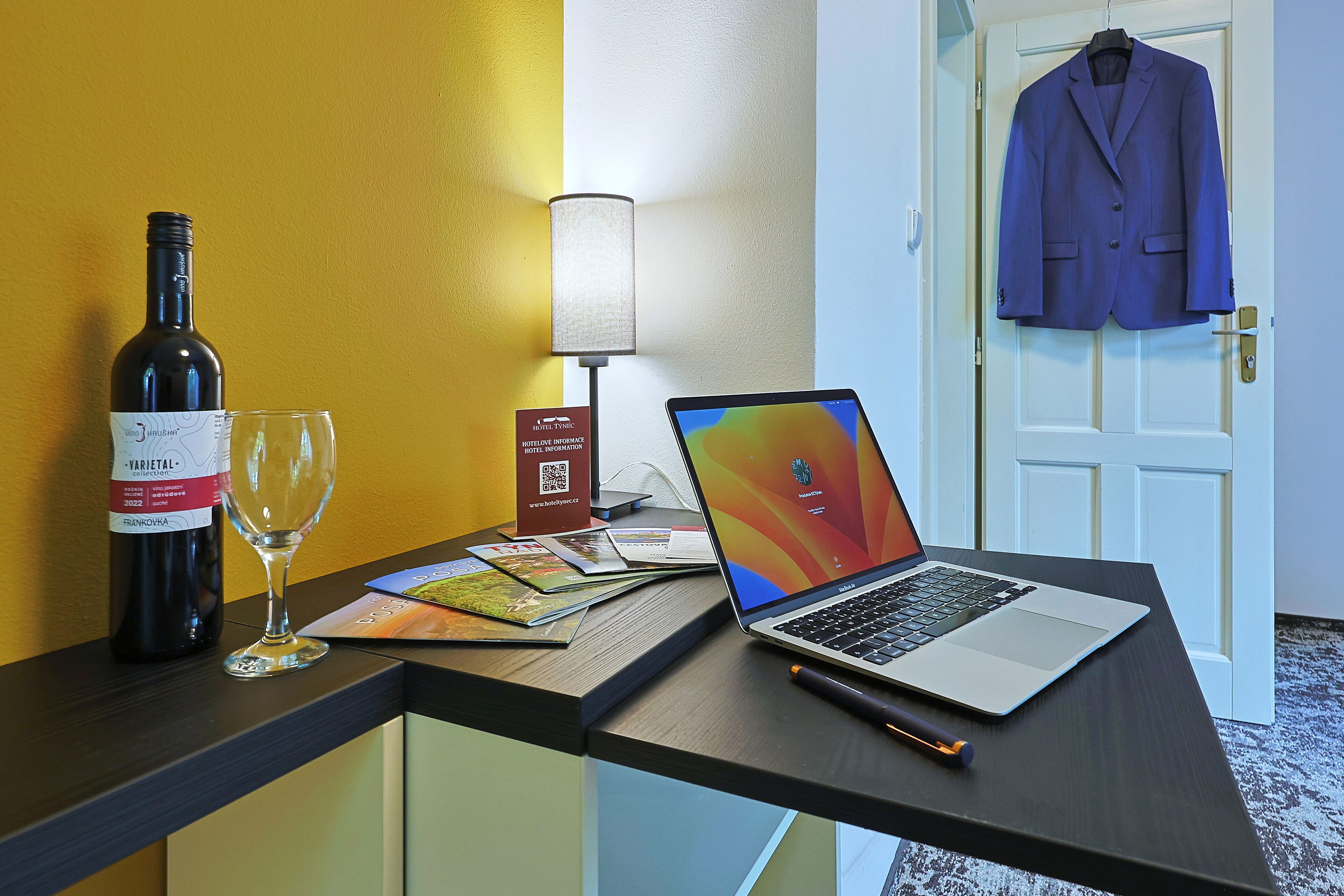 Un bureau avec un ordinateur portable, une bouteille de vin et un verre. Un manteau est accroché à la porte.