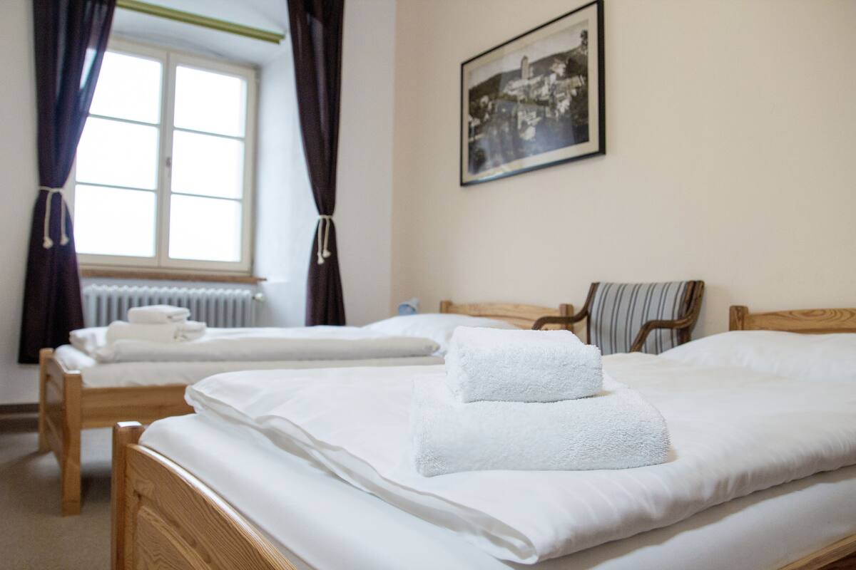 Habitación de hotel con dos camas de madera, ropa de cama blanca, una ventana con cortinas, un radiador, un cuadro en la pared y una silla a rayas.