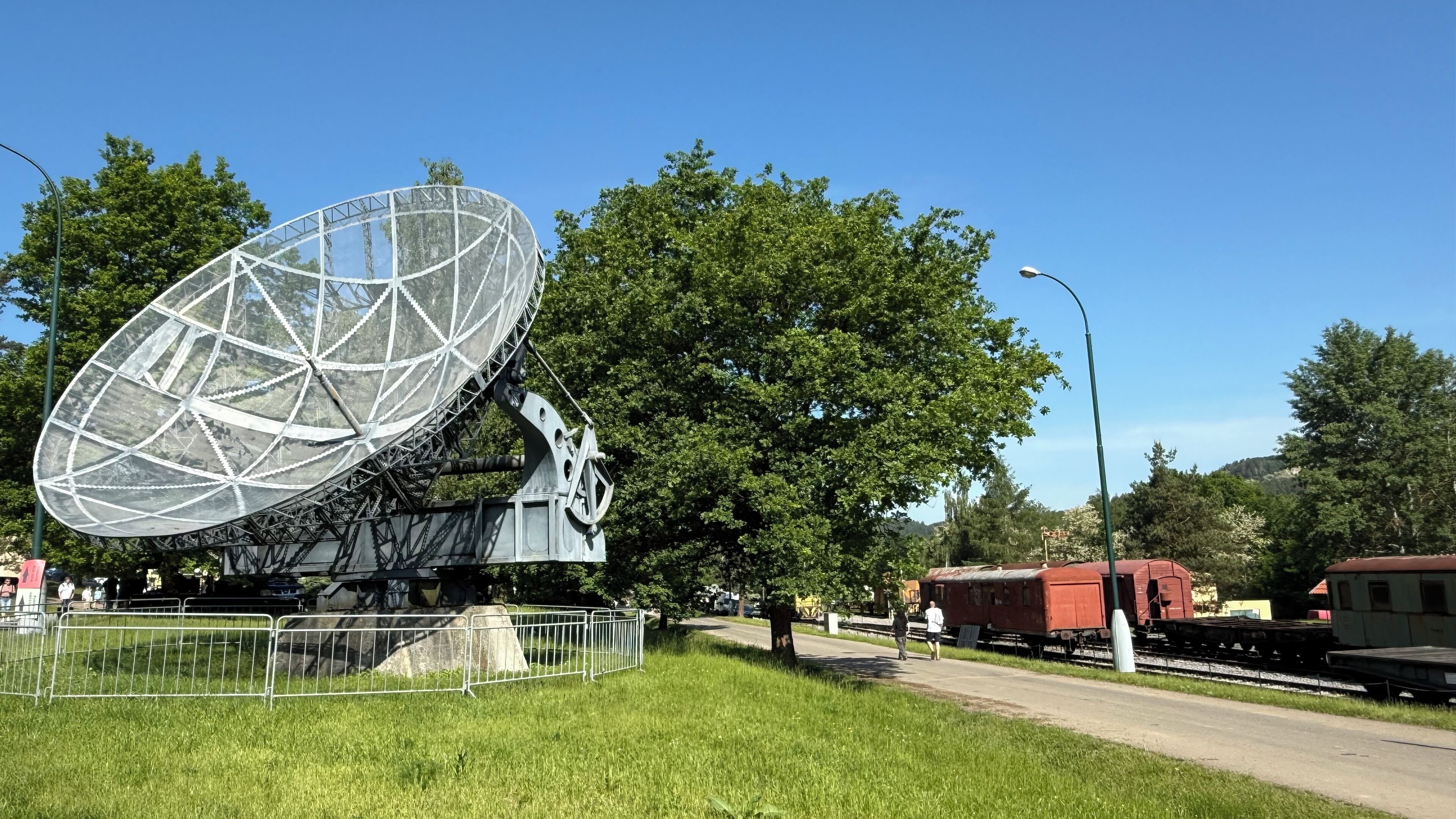 Una gran antena parabólica, un tren rojo sobre rieles y un cielo azul despejado.