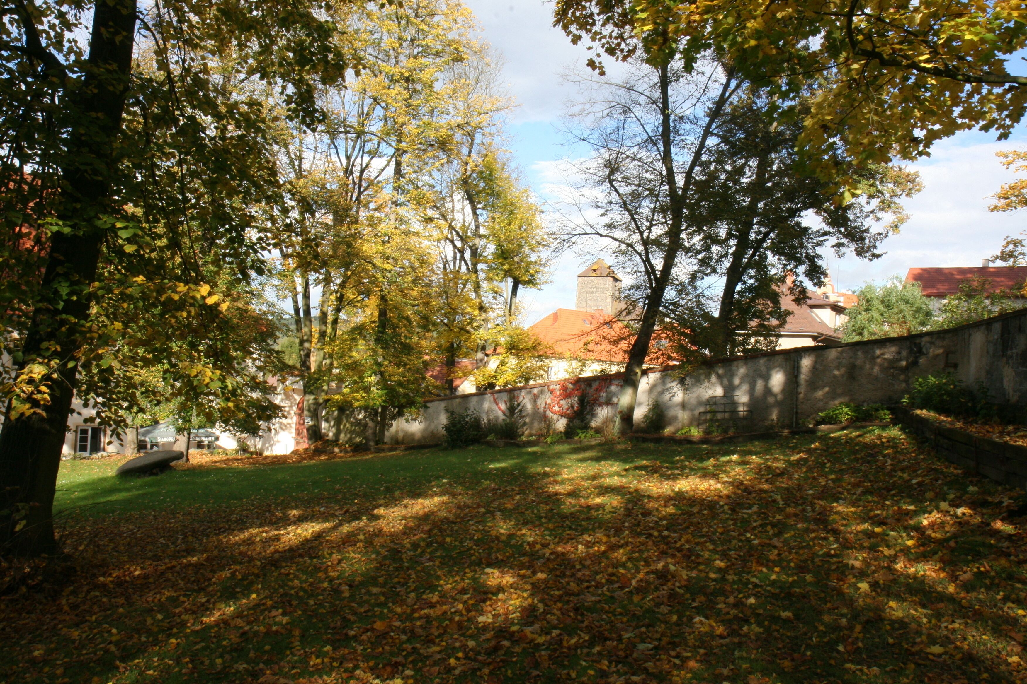 A courtyard with trees, a stone wall, and a building with a red roof in the background.