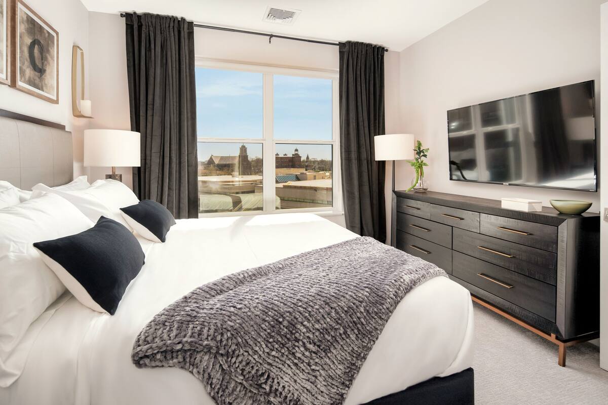 Une chambre d’hôtel moderne dotée d’un grand lit, d’un téléviseur à écran plat, d’une commode en bois foncé et offrant une vue sur le paysage urbain grâce à de grandes fenêtres.