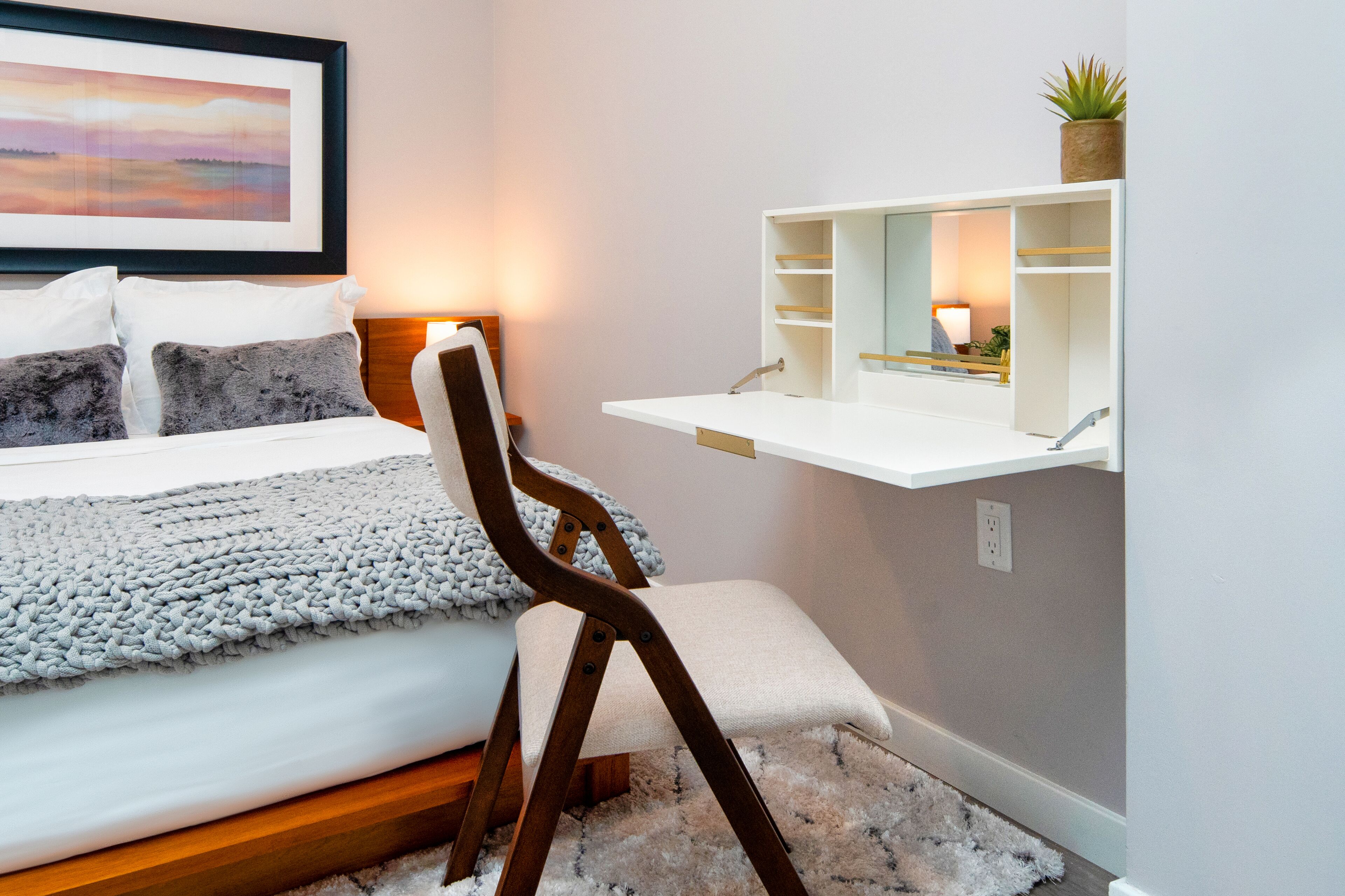 A bedroom with a bed, a chair, a bedside table with a mirror, and a shelf with a plant.