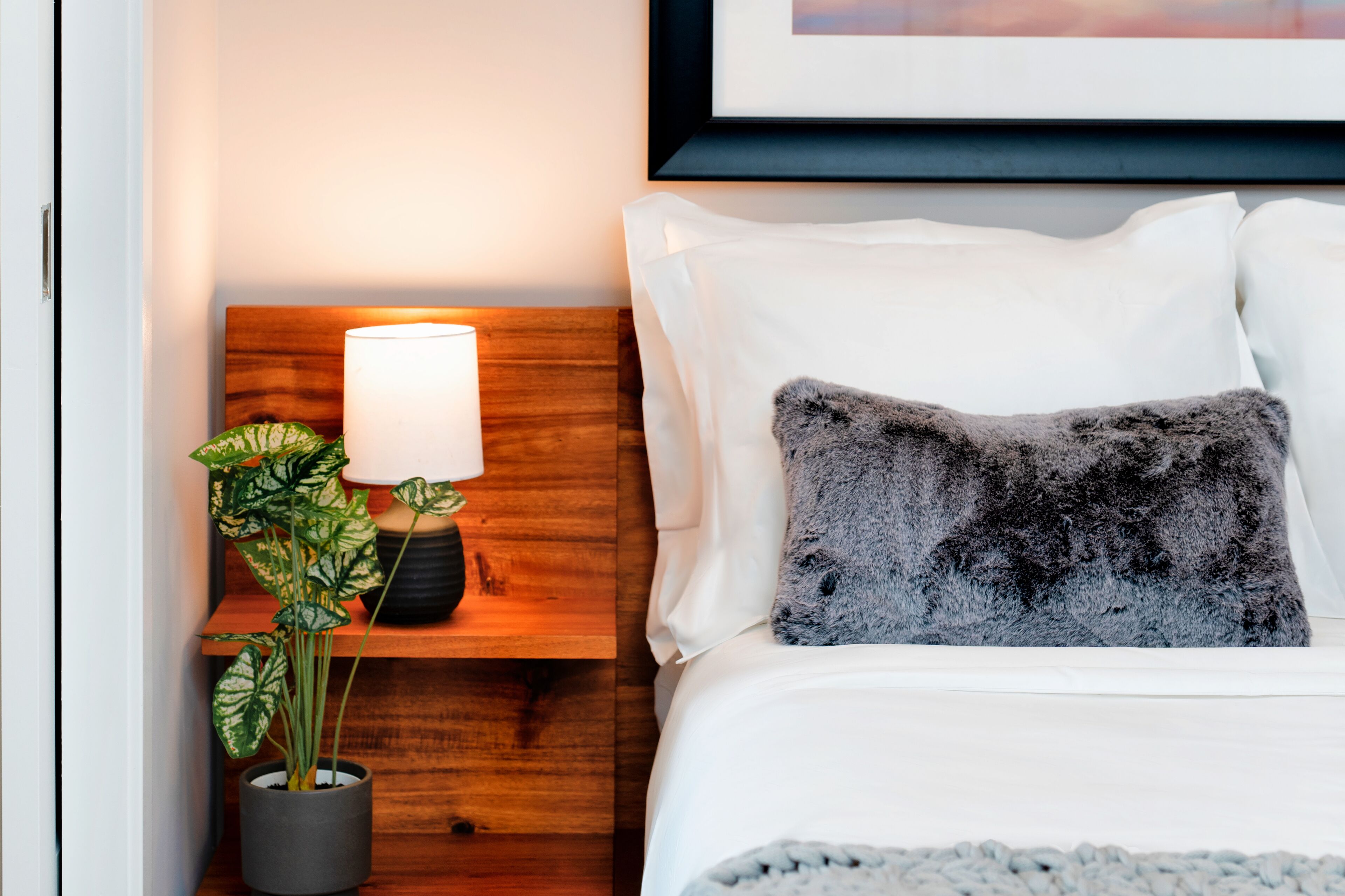 Una cama bien hecha con sábanas blancas y un cojín gris oscuro, una lámpara de noche con pantalla blanca y una mesita de noche de madera con una planta en maceta.