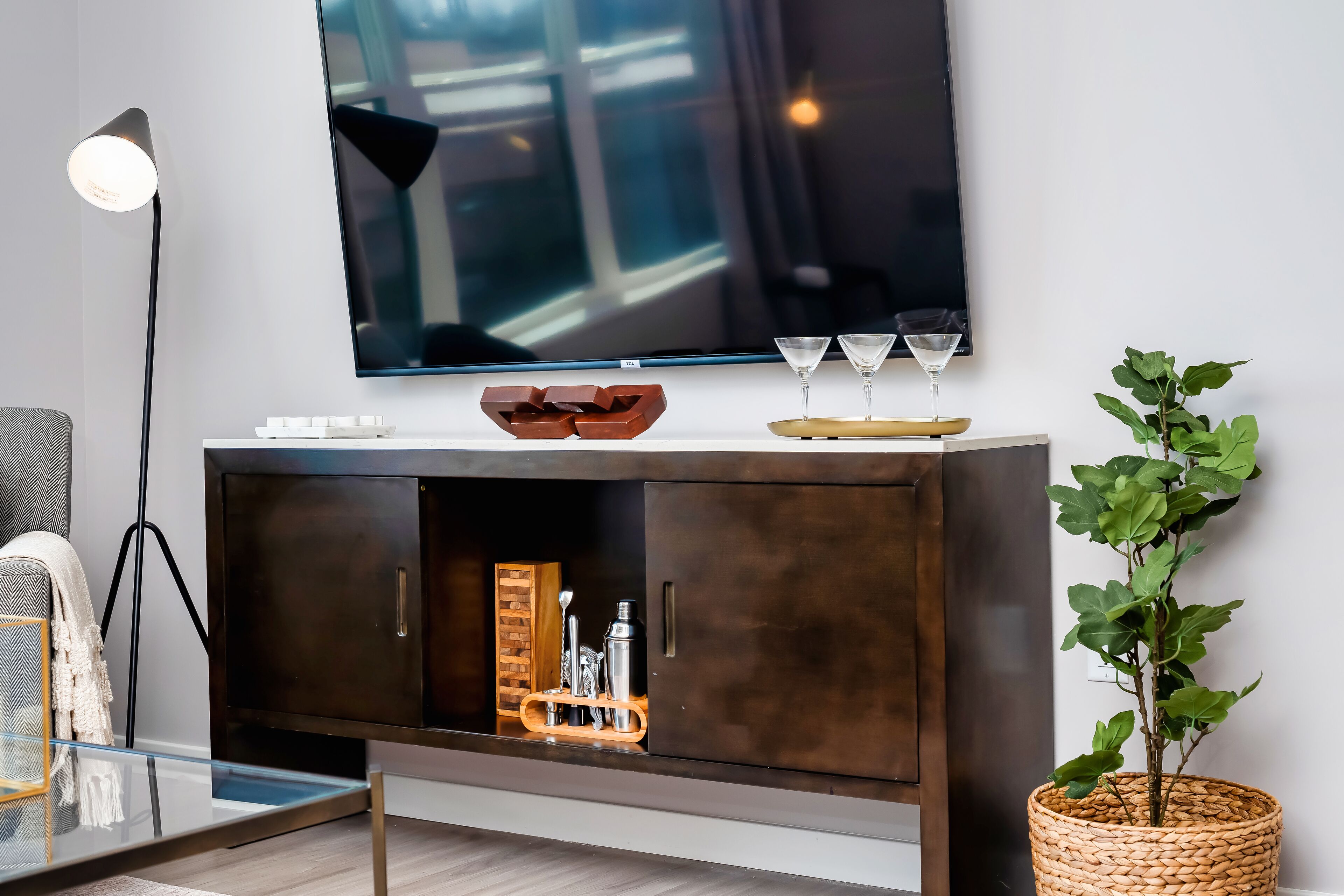 Un salon moderne avec un téléviseur à écran plat fixé au mur, un meuble en bois foncé doté d’une étagère en verre et une plante en pot.
