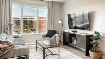A modern living room with a sofa, a glass coffee table, a TV, and a large window with a view of buildings.