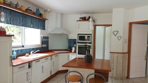 A compact kitchen with white cabinets, a wooden countertop, a built-in oven, a microwave, and a sink. There are two bar stools and a potted plant on the island. - Bassin d'Arcachon family home with swimming pool (LE TEICH)