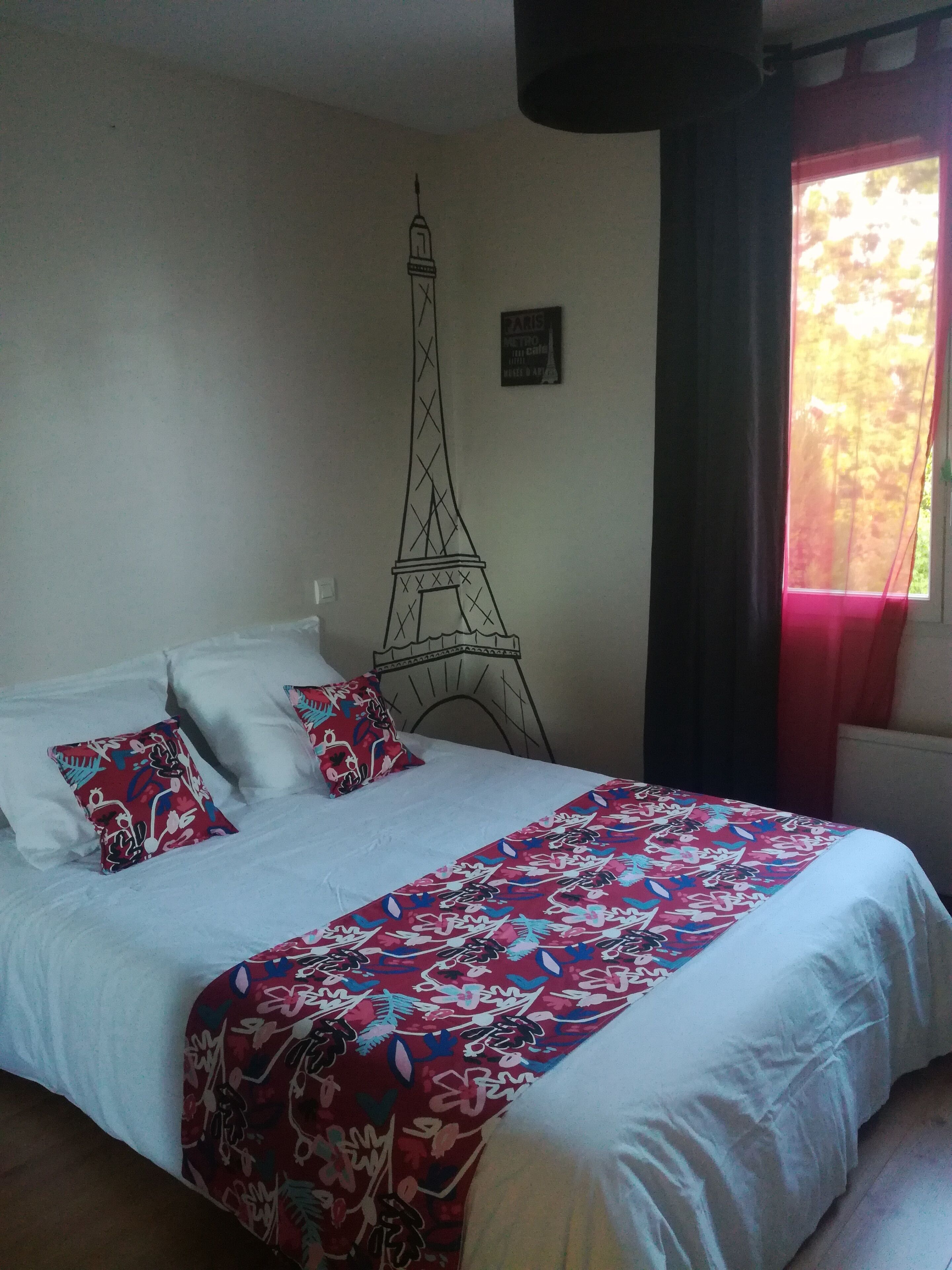 Een bed met witte lakens en een rode sprei met bloemenprint, een muur met een tekening van de Eiffeltoren, en een raam met rode gordijnen.