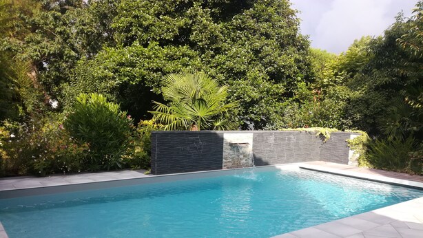 A rectangular swimming pool with a waterfall feature, surrounded by lush greenery and a tiled deck. - Bassin d'Arcachon family home with swimming pool (LE TEICH)