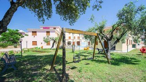 A playground with swings and a bench, surrounded by trees and a building with a pink facade.