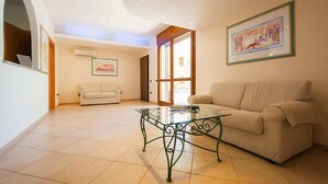 A spacious living room with a glass coffee table, two sofas, a painting on the wall, and a door leading outside.