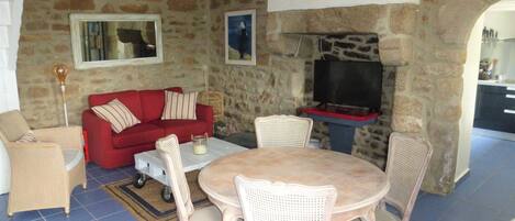 A cozy living room with a stone wall, a red sofa, wicker chairs, a wooden table, and a television.