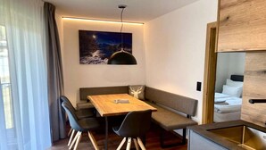 A modern room with a wooden dining table, bench seating, a kitchen area, and a large window with curtains. - Bergzeit Apartments near ski slope (Saalbach-Hinterglemm)