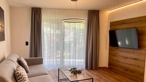 A modern living room with a sofa, a coffee table, a TV, and a large window with curtains. - Bergzeit Apartments near ski slope (Saalbach-Hinterglemm)