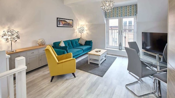 Una sala de estar moderna con un sofá color turquesa, un sillón amarillo, una mesa de centro blanca, un televisor y una mesa de comedor de vidrio.